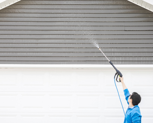 Man in blue jacket cleaning exterior siding under the roof by high pressure nozzle spray